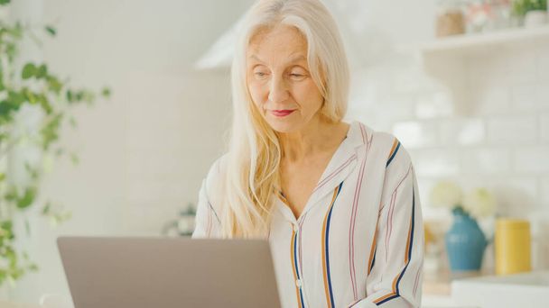 Beautiful Senior Woman Using Laptop Computer in a Sunny Kitchen. She Smiles. Grandmother is Connected with Children via internet and Social Media. Pensioner Working from Home. - 写真・画像