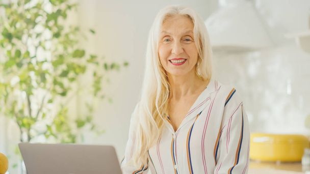 Beautiful Senior Woman Using Laptop Computer in a Sunny Kitchen. She Looks at Camera and Smiles. Grandmother is Connected with Children via internet and Social Media. Pensioner Working from Home. - Zdjęcie, obraz