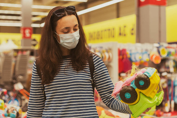 La mujer con la máscara protectora en el supermercado escoge los juguetes infantiles. Mujer en máscara médica en el supermercado tonificado. - Foto, imagen