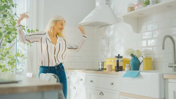Authentic Senior Female in Blue Jeans and White Striped Blouse is Creatively Dancing in the Kitchen. Sunny Modern Kitchen with Healthy Lifestyle Vibes. Happy Beautiful Pensioner Relaxing at Home. - Fotoğraf, Görsel