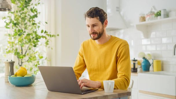 Young Handsome Man Using Laptop Computer and Drinking Morning Cup of Coffee or Tea While Wearing a Yellow Pullover. Curly-hared Male Sitting in a Modern Sunny Kitchen. Freelancer Working from Home. - Zdjęcie, obraz