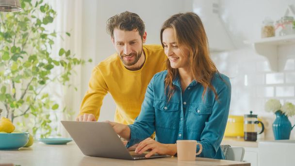 Beautiful Young Couple Using Laptop Computer in Kitchen at Home. Female Working on a Project. Boyfriend is Helping Girlfriend with Online Work. Female Browsing Internet and Doing Online Shopping. - Foto, Imagem