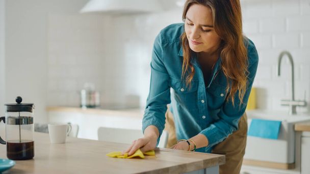 Caring Housewife Wiping Spilled Coffee or Crumbs from a Wooden Kitchen Table. Beautiful Young Female Using a Household Cleaner Wipe for Cleaning. Brunette is Wearing a Jeans Shirt and Beige Pants. - Photo, Image