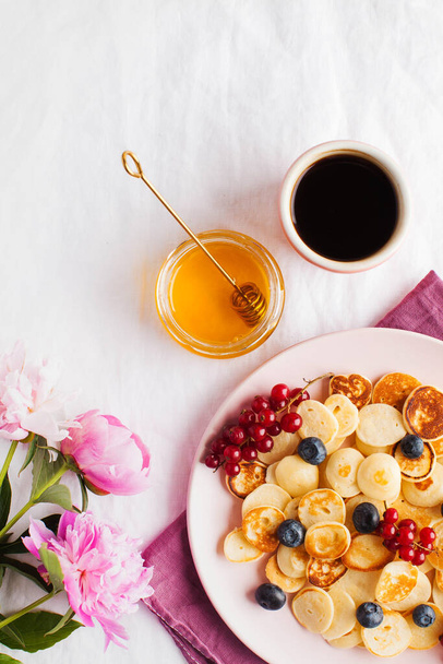 Tiny pancakes with berries, honey and flowers on a white tablecloth and space for text. Pancake cereal. The concept of Breakfast, food trends. Copy space. . High quality photo - Фото, изображение