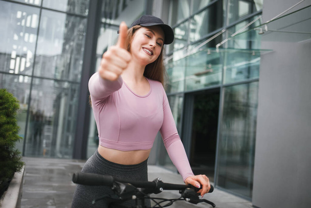 Lány sapkában és sportruházat mutatja hüvelykujját, miközben ül a kerékpáron - Fotó, kép