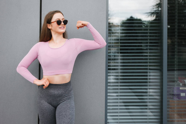 Mujer atlética positiva en gafas de sol muestra bíceps
 - Foto, Imagen