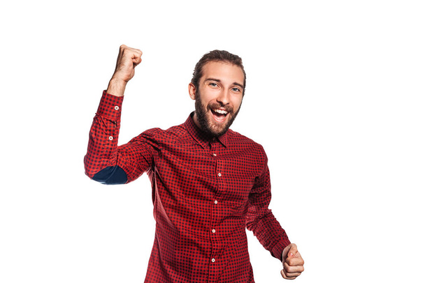 Young man with a beard in a red shirt rejoices with hands up on a white background, isolate - Fotó, kép
