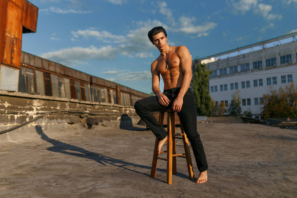 Bonito jovem musculoso e apto esportivo com tronco nu, vestido com jeans pretos, posando na velha fábrica abandonada. Visão de comprimento total
. - Foto, Imagem