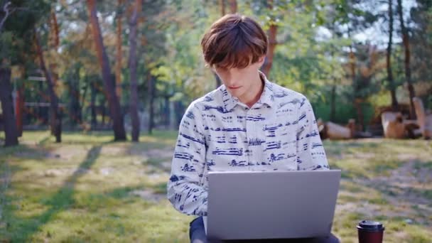 In a beautiful large park student guy using a laptop to studying at fresh air he take coffee to get more concentrated - Záběry, video