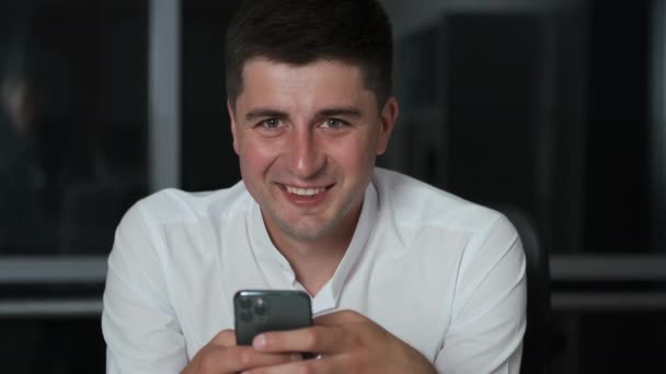 Cute young man with a Caucasian appearance lingered in the office at work until night. Sits on the phone in social networks and smiles - Materiał filmowy, wideo
