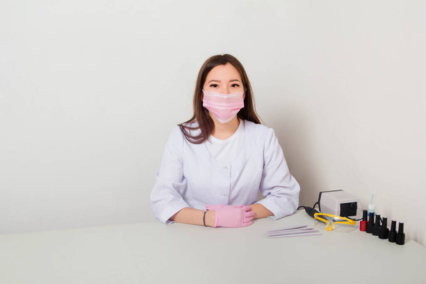A woman in a white coat with a mask on her face sits at a table with spread out manicure tools. Studio photo on a white background. - Foto, immagini