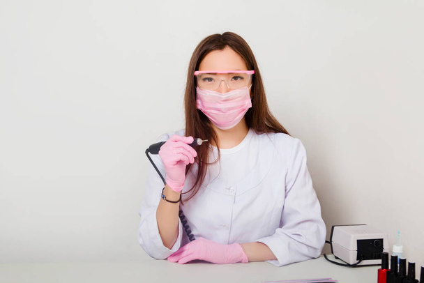 A woman is a master of nails, in a white coat with a mask and glasses on her face, sits at a table with spread out manicure tools and holds a manicure machine in her hands. Photo on a white background. - Foto, afbeelding