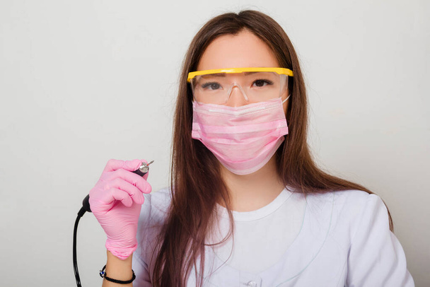 Portrait of a nail master woman in a white coat, gloves, mask and glasses on her face, standing on a white background with a manicure machine in her hands. - Foto, Imagem