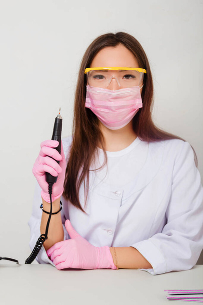 Portrait of a nail master woman in a white coat, gloves, mask and glasses on her face, standing on a white background with a manicure machine in her hands. - Foto, Imagen