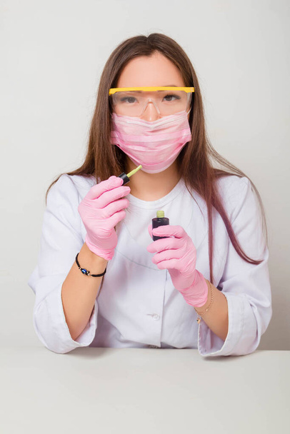 A woman in gloves and a white coat with a mask and glasses on her face sits at a table and holds nail polishes in her hands. Studio photo on a white background. - Photo, Image