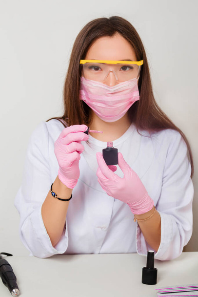 A woman in gloves and a white coat with a mask and glasses on her face sits at a table and holds nail polishes in her hands. Studio photo on a white background. - Foto, Imagen