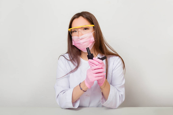 A woman in gloves and a white coat with a mask and glasses on her face sits at a table and holds nail polishes in her hands. Studio photo on a white background. - Foto, Imagem