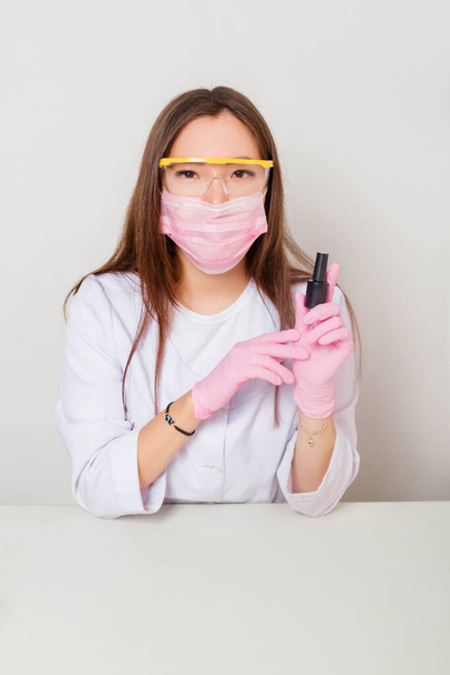 A woman in gloves and a white coat with a mask and glasses on her face sits at a table and holds nail polishes in her hands. Studio photo on a white background. - Фото, изображение