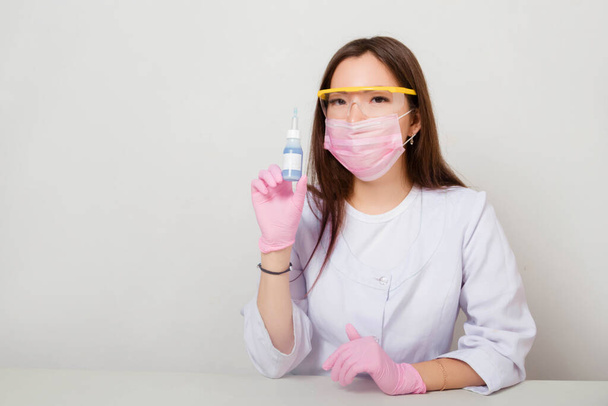 A woman in gloves and a white coat with a mask and glasses on her face sits at a table and holds a bottle of nail polish in her hands. Studio photo on a white background. - Fotoğraf, Görsel