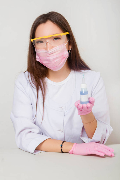 A woman in gloves and a white coat with a mask and glasses on her face sits at a table and holds a bottle of nail polish in her hands. Studio photo on a white background. - Foto, Bild