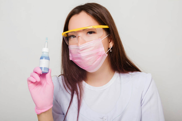 A woman in gloves and a white coat with a mask and glasses on her face sits at a table and holds a bottle of nail polish in her hands. Studio photo on a white background. - Foto, Bild