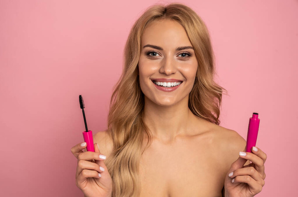 Beauty portrait of a lovely young woman with long blonde hair standing isolated over pink background, showing eye mascara. - Foto, immagini