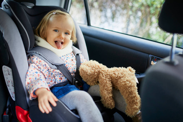 Adorable toddler girl in modern car seat with her favorite stuffed toy. Little kid traveling by car. Child safety on the road. Trip with a baby - Foto, Imagen