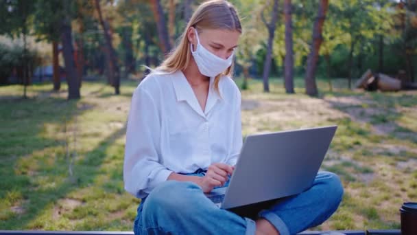 Good looking lady with a protective mask sitting on the chair in park she take her laptop to working online in the park while is pandemic of Coronavirus - Materiał filmowy, wideo