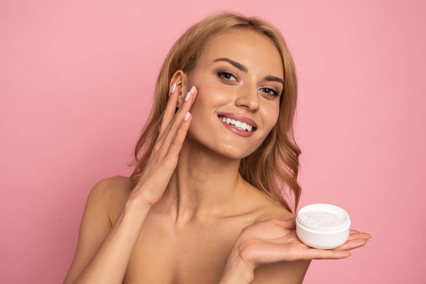 Close up portrait of happy gorgeous smiling attractive satisfied young woman with beaming smile, she is holding a cream jar and touching her cheek, isolated on pink background. - Foto, Imagen