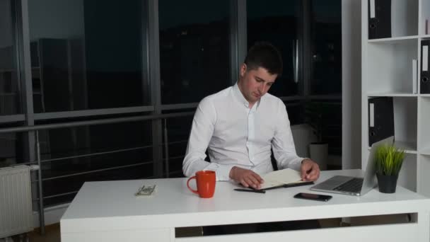 The 30-year-old director of the company sits in the evening office until late drinking coffee or tea and reading a book. Panorama of night city from the office - Séquence, vidéo