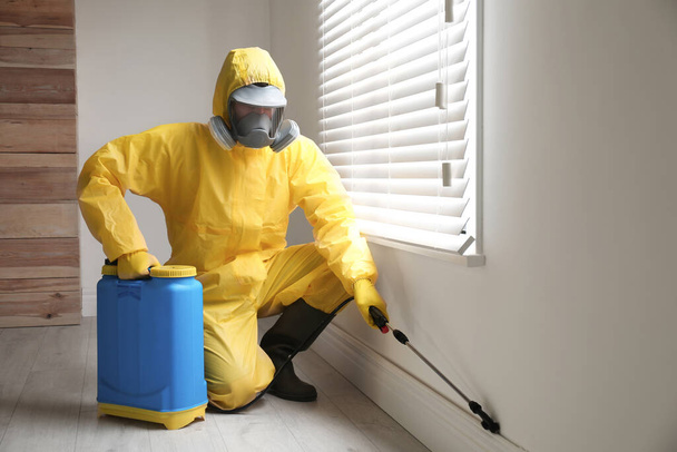 Pest control worker in protective suit spraying pesticide near window indoors - Foto, afbeelding