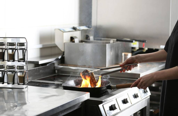 Female chef cooking meat with burning flame on stove in restaurant kitchen, closeup - Photo, image