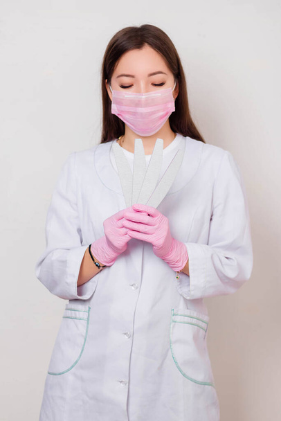 Portrait of a woman master of nails, in a white coat and pink mask on her face with nail files in her hands. Photo on a white background. - Фото, изображение
