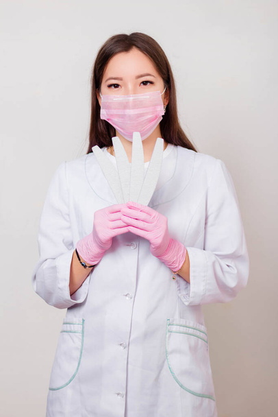 Portrait of a woman master of nails, in a white coat and pink mask on her face with nail files in her hands. Photo on a white background. - Foto, imagen