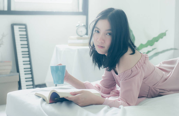 Itseni vuoksi. Mukavuutta ja rentoutumista. Kaunis nuori aasialainen nainen juo teetä tai kahvia ja lukee kirjaa nukkuessaan sängyssä kotona - Valokuva, kuva