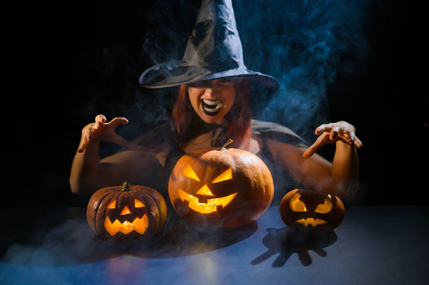 An ominous witch in a hat conjures over a jack-o-lantern. Traditional halloween characters. Mystical fog creeps over pumpkins with carved terrible faces. Woman in carnival costume. - Foto, afbeelding