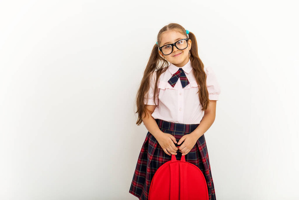 Teljes hosszúságú portré mosolygós iskolás lány egyenruhában és szemüveg, kezében piros hátizsák álló fehér háttér - Fotó, kép