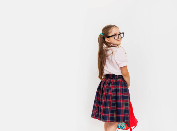 Teljes hosszúságú portré mosolygó iskolás lányról egyenruhában és szemüveg, kezében piros hátizsák áll fehér háttér másolási hely szöveg - Fotó, kép