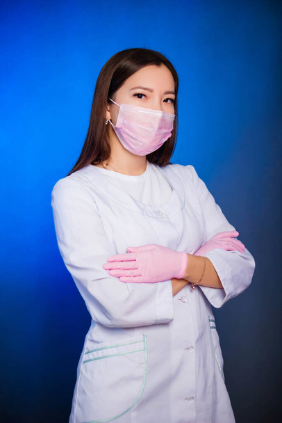Portrait of a woman in a white coat and mask with hands in gloves folded over her chest. Photo on a blue background. - Foto, immagini
