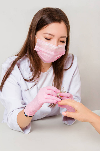 The woman is a manicurist in a white coat, mask and gloves, paints a clients nails. - Fotó, kép