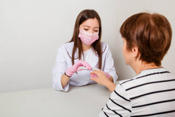 The woman is a manicurist in a white coat, mask and gloves polishes her nails with a manicure machine to a client. - 写真・画像