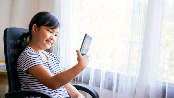 Asian little girl using smartphone for video call on a chair at home - Photo, Image