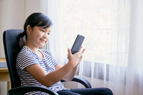Азійська дівчинка користується смартфоном для відеодзвінків на стільці вдома. - Фото, зображення