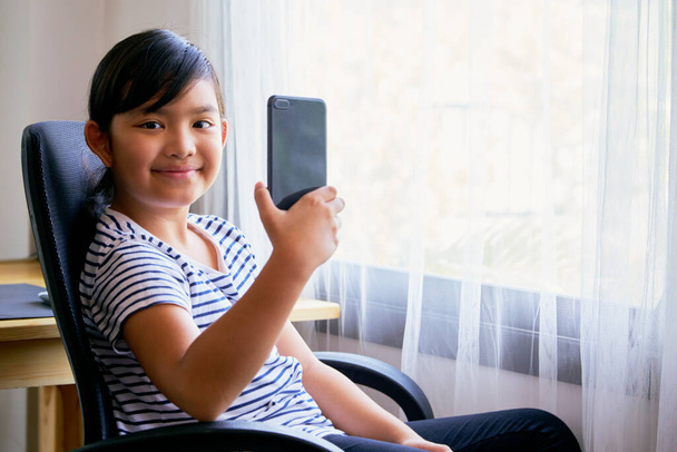 Aasialainen pieni tyttö, joka käyttää älypuhelinta videopuheluun tuolilla kotona - Valokuva, kuva