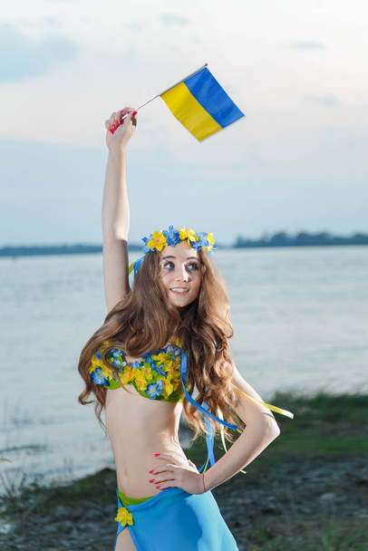Dziewczyna w niebiesko-żółtym stroju kąpielowym z kwiatami, ukraińską flagą i wieńcem ze wstążkami na głowie, stojąca nad brzegiem rzeki. Ukraina narodowe kolory, odzież i akcesoria dla kobiety. Niezależna Ukraina, koncepcja wolności. - Zdjęcie, obraz