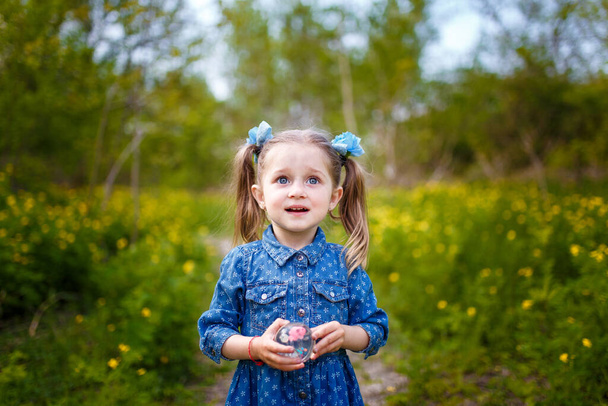 Grappig klein meisje met twee paardenstaarten in blauwe jurk tussen bloeiende gele bloemen van celandine. Voorjaarsbloesem en zorgeloos kinderconcept. - Foto, afbeelding