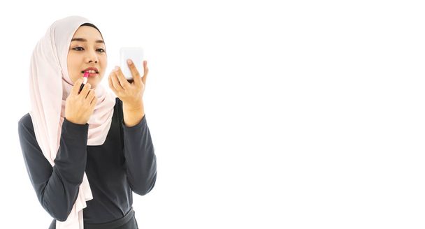 Retrato de una joven musulmana joven y sonriente usando Hijab y pintándose los labios con un lápiz labial con espejo sobre fondo blanco tomado en el estudio. Usar para el concepto de moda y belleza
 - Foto, Imagen