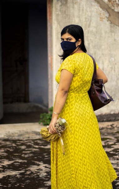 una mujer india en vestido amarillo y mascarilla está saliendo con paraguas y bolsa después de desbloquear 1.0 de pandemia de virus de la corona
 - Foto, imagen