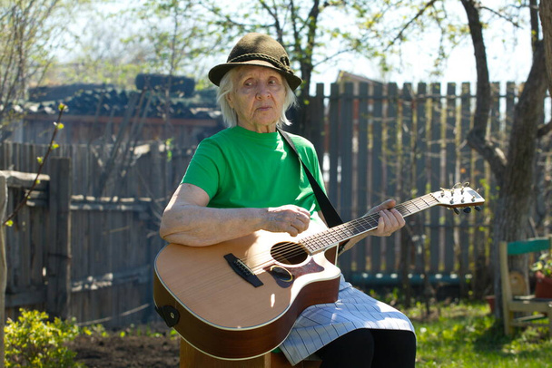 Portrait of a grandmother in nature. Grandmother with an acoustic guitar in a green T-shirt and a cap in the spring garden. - Photo, Image