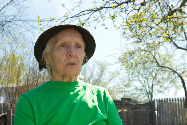 Portret van een grootmoeder in de natuur. Grootmoeder in een groen T-shirt en een pet in de tuin in het voorjaar. Gezicht dicht. - Foto, afbeelding